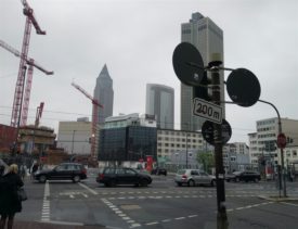 Stadtansicht. 4-teilige Kreuzung, Verkehrsschilder im Vordergrund, Passanten, im Hintergrund mehrere Hochhäuser, eines davon im Bau