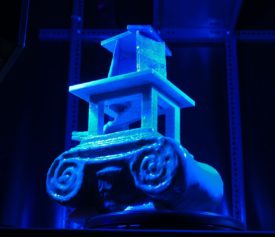 3D-Druck-Objekt. als Basis der Kopf einer anktiken Säule, darauf ein Konstrukt, in dem eine Wendeltreppe nach oben führt in eine Art Turm. auf einem Drehteller unter violettem und blauen Licht.