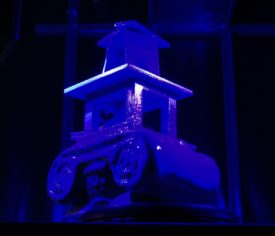 3D-Druck-Objekt. als Basis der Kopf einer anktiken Säule, darauf ein Konstrukt, in dem eine Wendeltreppe nach oben führt in eine Art Turm. auf einem Drehteller unter violettem und blauen Licht.