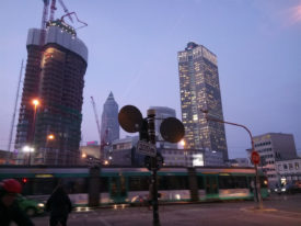 Stadtansicht. 4-teilige Kreuzung, Verkehrsschilder im Vordergrund, Passanten, im Hintergrund mehrere Hochhäuser, eines davon im Bau